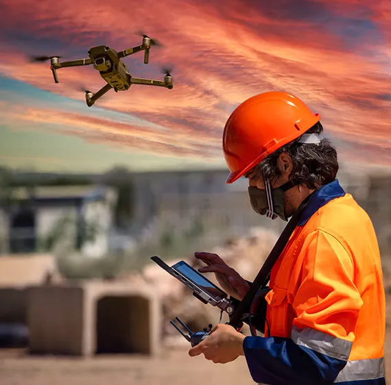 Drone pilot in foreground with orange safety hat and shirt with drone flying in the background.