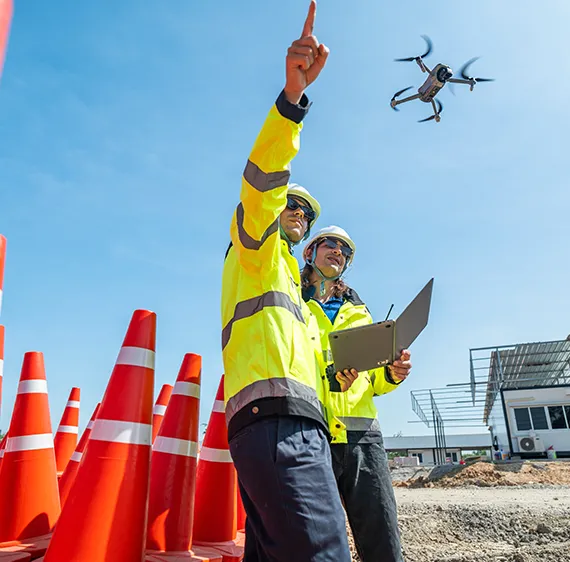 2 drone operators in safety vests near orange construction cones pointing in the air with a drone flying overhead.