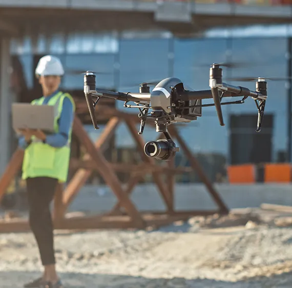 Drone flying in foreground on construction site with operator holding laptop in the background.